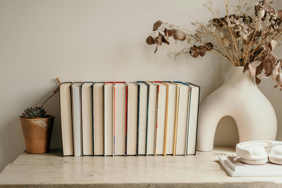 Row of books next to a small potted plant and a white vase with dried flowers on a wooden surface.