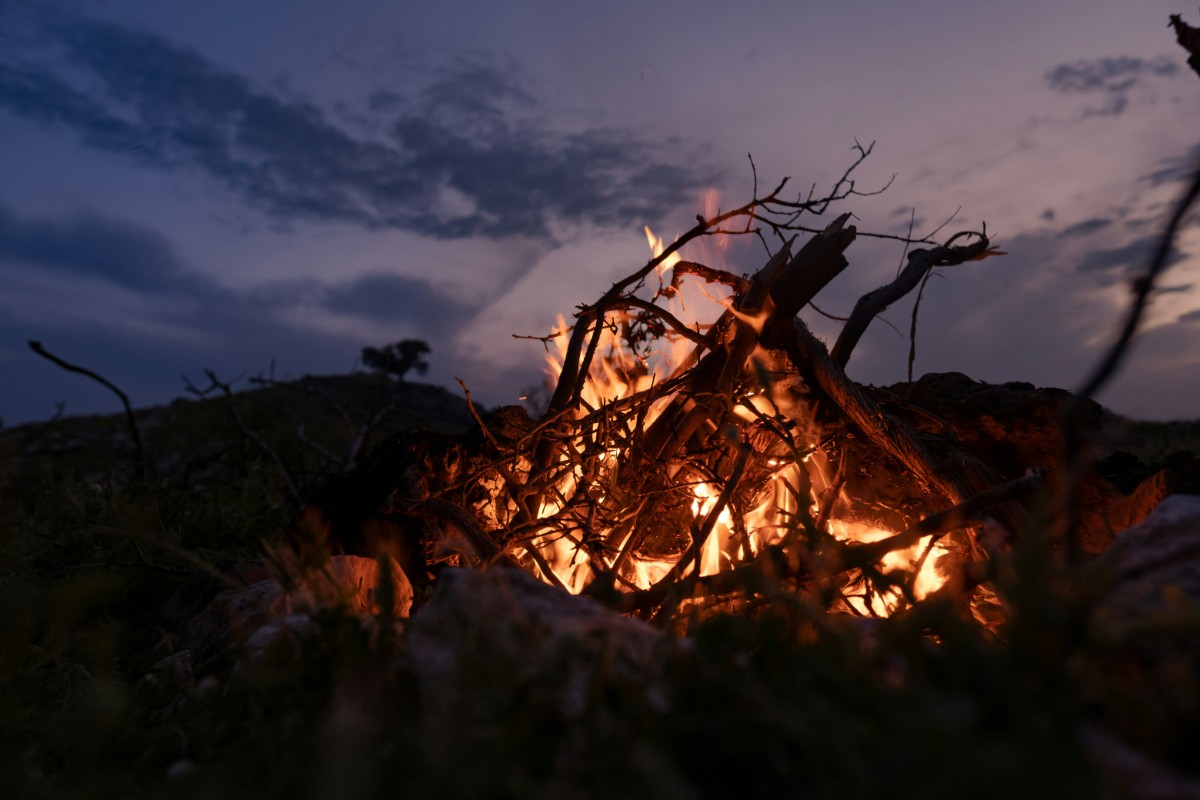 A campfire burns with bright flames and branches under a darkening twilight sky.