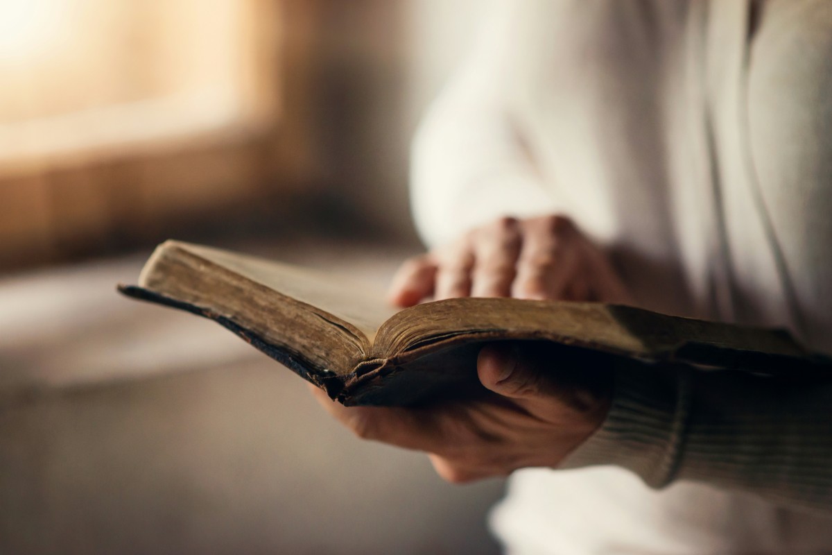 A person holding and reading an open book with one hand, while the other hand points at a page.