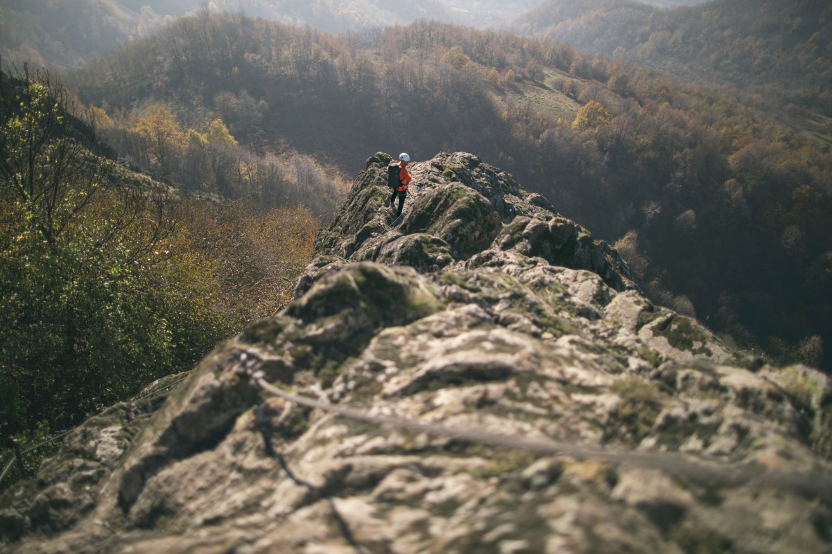 A person wearing a red jacket stands on a rocky ridge with forested hills in the background.