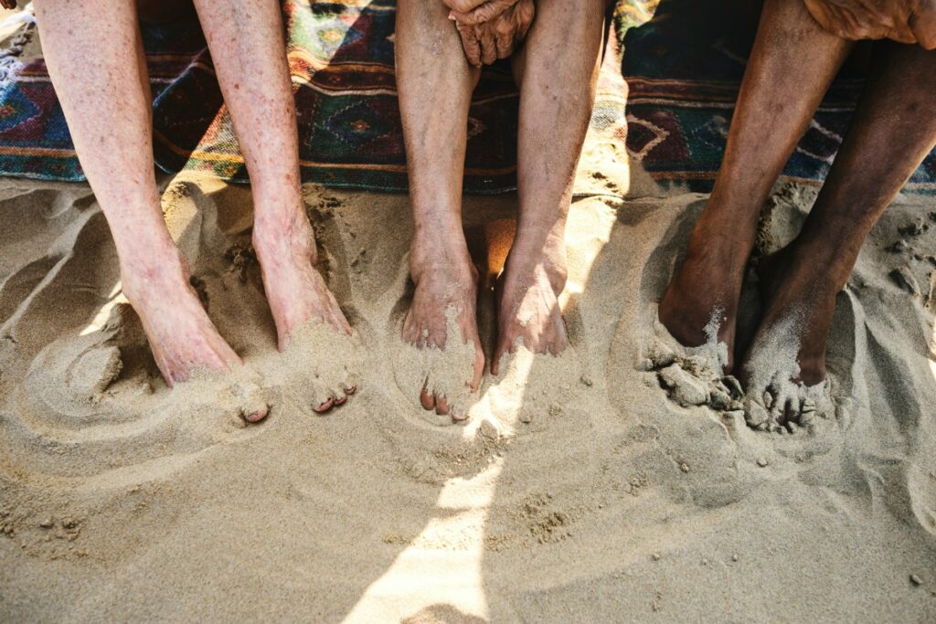 Three pairs of legs partially covered with sand, resting on a beach blanket at the shore.