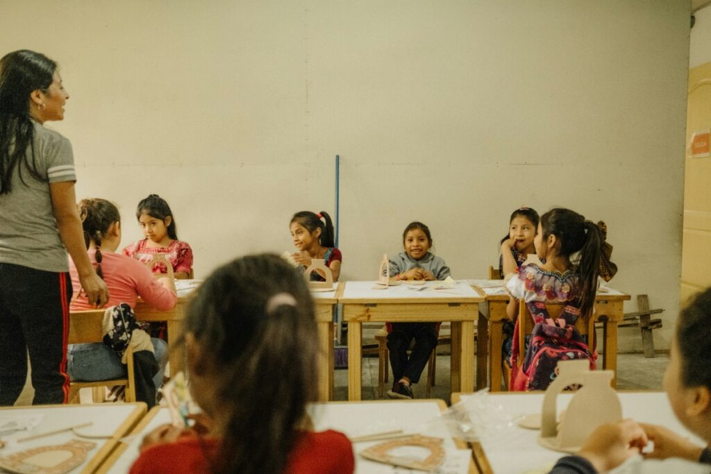 A group of children sitting at desks in a classroom, with a teacher standing at the front. Some children appear to be talking, and art supplies are on the tables.