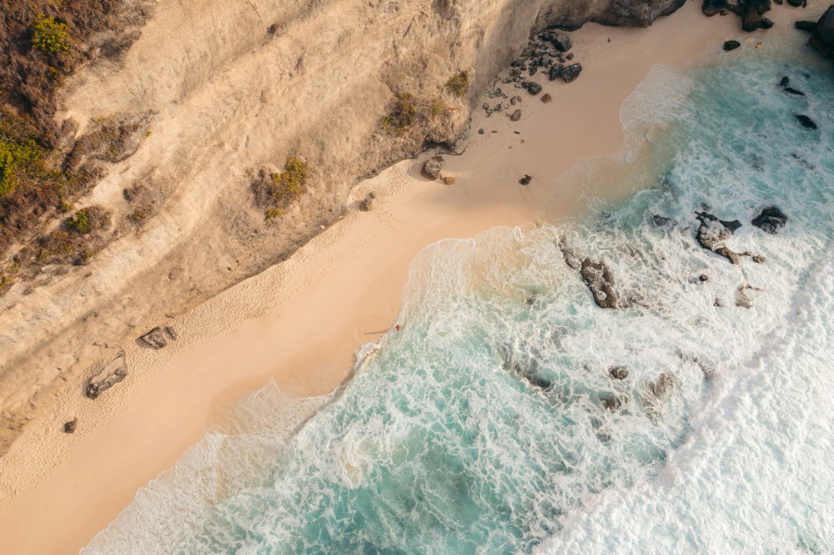 Aerial view of ocean waves crashing on a sandy beach with a steep cliff on the left side.