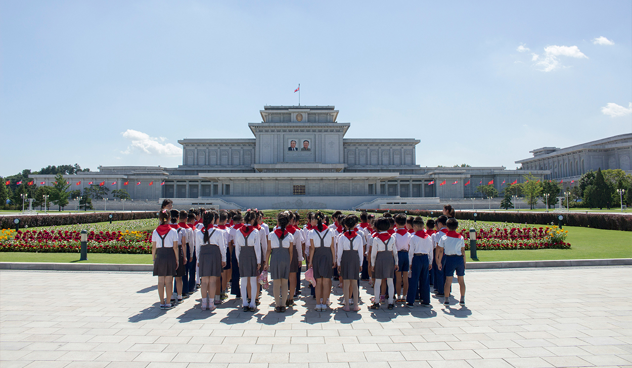 A group of children in uniforms standing in from of a grass block with flowers.