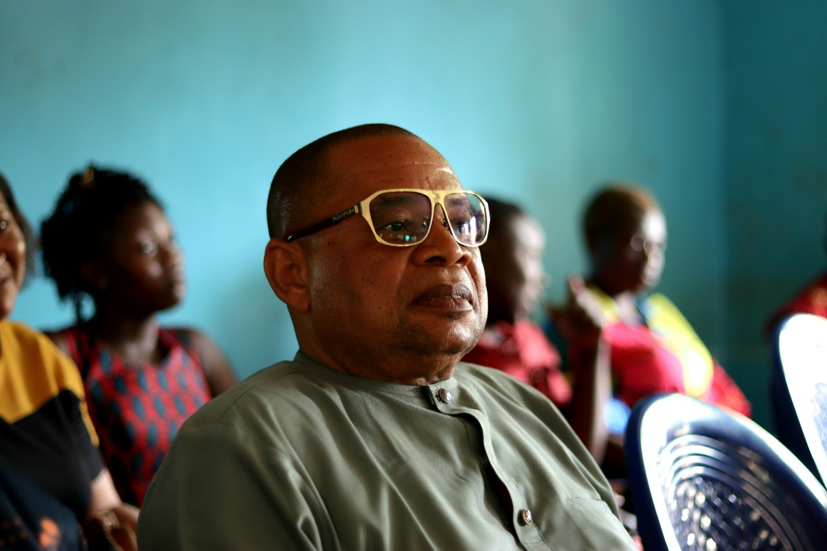 A man wearing glasses and a green shirt sits on a plastic chair in a room, with other people seated in the background.