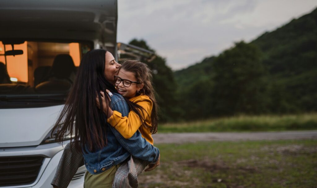 A woman smiles as she holds a child near a camper van in a grassy area with trees in the background.