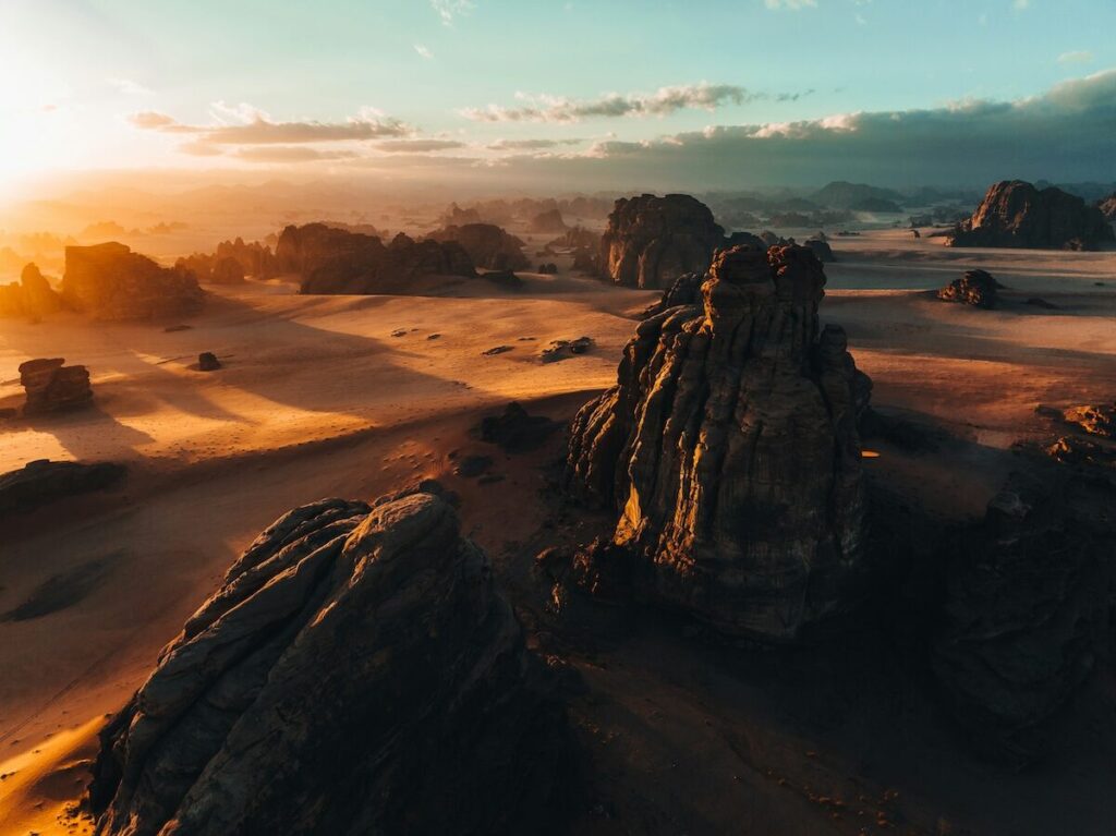 Rock formations stand in a vast, sandy desert landscape at sunset, with long shadows cast and a partly cloudy sky above.