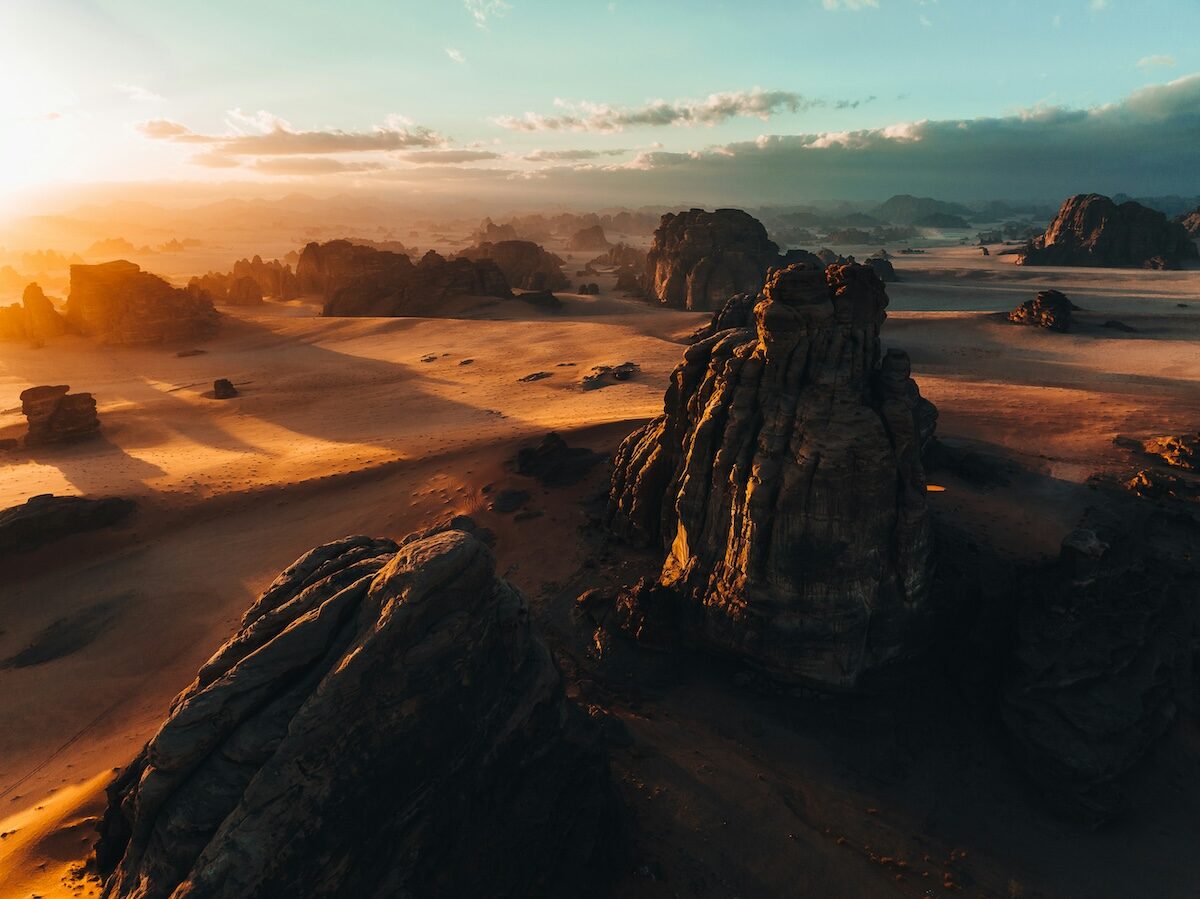 Rock formations stand in a vast, sandy desert landscape at sunset, with long shadows cast and a partly cloudy sky above.