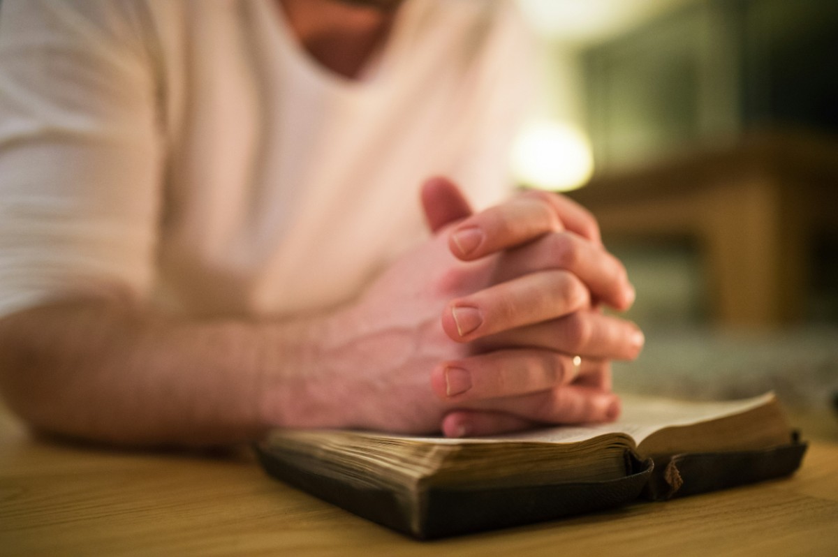 Person in a white shirt with hands clasped on an open book, possibly a Bible, on a wooden table.