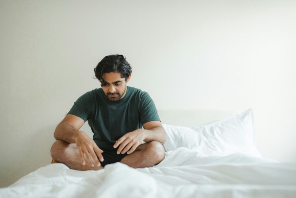 A man sits cross-legged on a bed with white sheets, looking down thoughtfully, wearing a green t-shirt and black shorts.