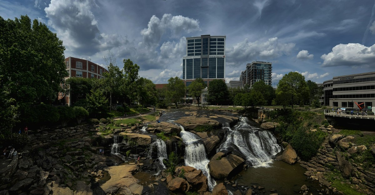Urban park scene with cascading waterfalls in the foreground and tall buildings in the background, under a partly cloudy sky.
