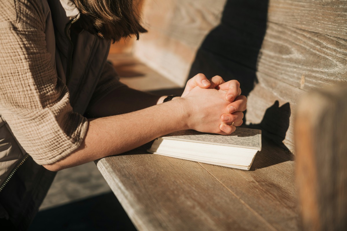 Person leaning over a bench with hands clasped, resting on an open book.