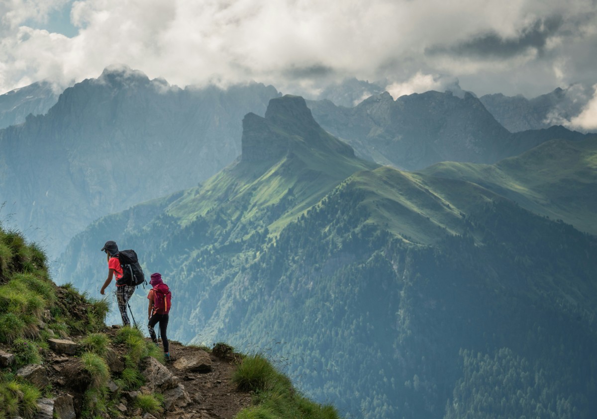 Two hikers walk along a rocky mountain trail with green peaks and clouds in the background.