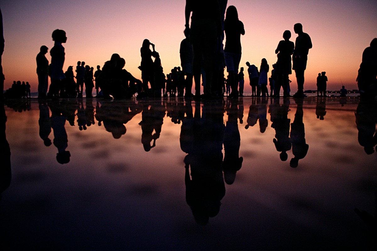 Silhouettes of people stand and interact on a reflective surface during sunset, creating mirrored reflections against a colorful sky.