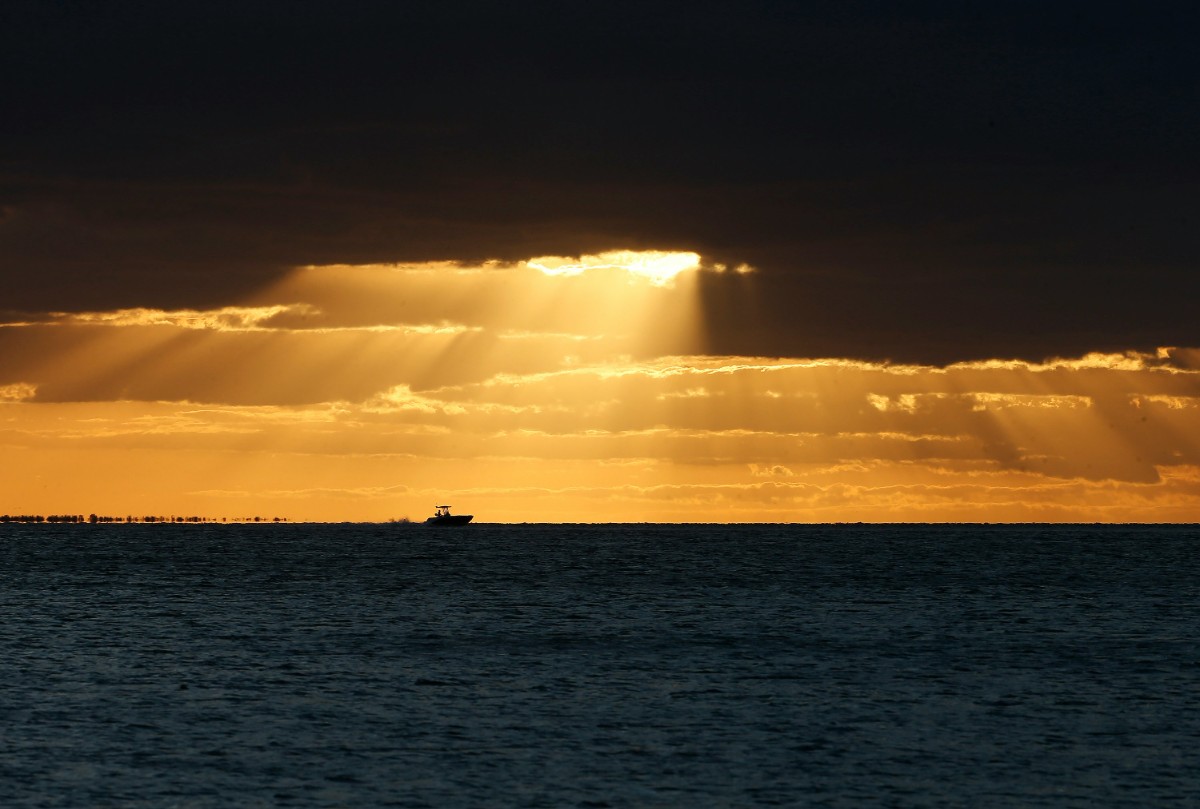 Sunlight breaks through clouds over a dark sea, illuminating the horizon with a warm glow, while a small boat is silhouetted against the bright sky.