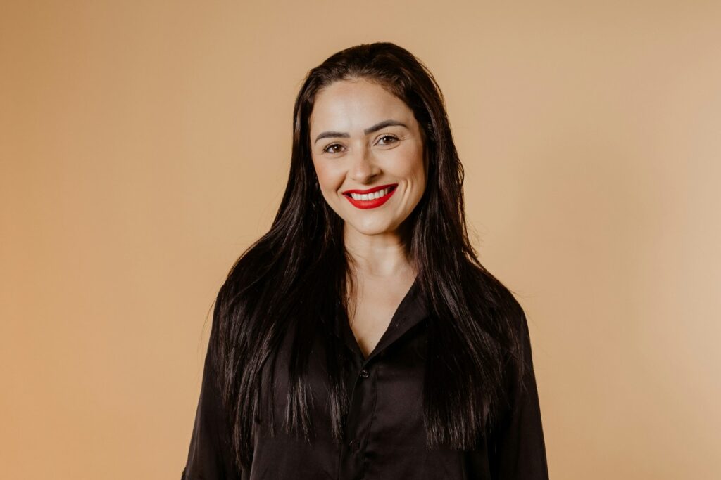 Woman with long dark hair and red lipstick, wearing a black blouse, smiles against a beige background.