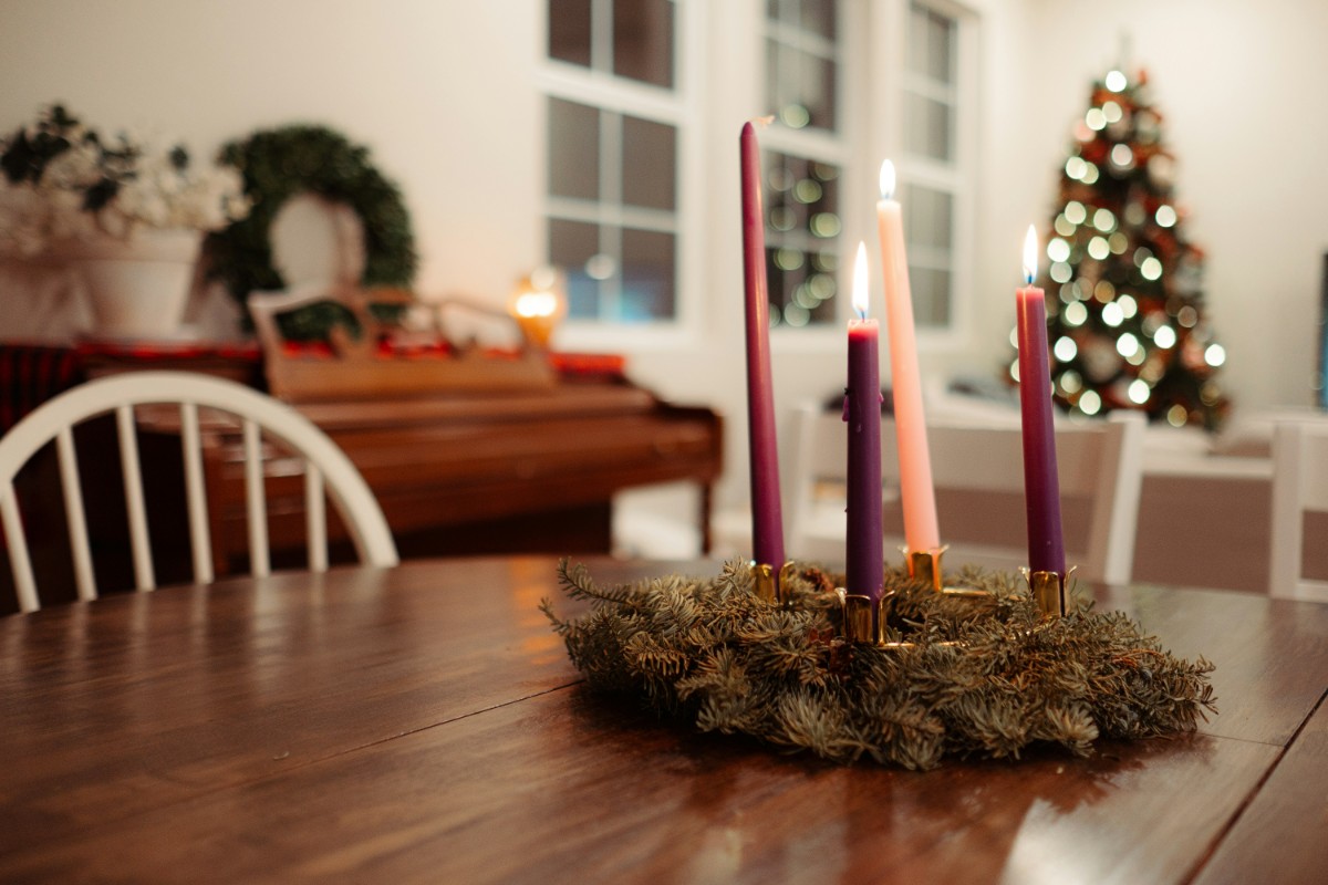 An Advent wreath with three purple candles and one pink candle is on a wooden table. A decorated Christmas tree and piano are in the softly lit background.