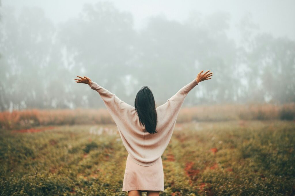 A person in a light dress stands in a field with arms outstretched, facing foggy trees in the distance.