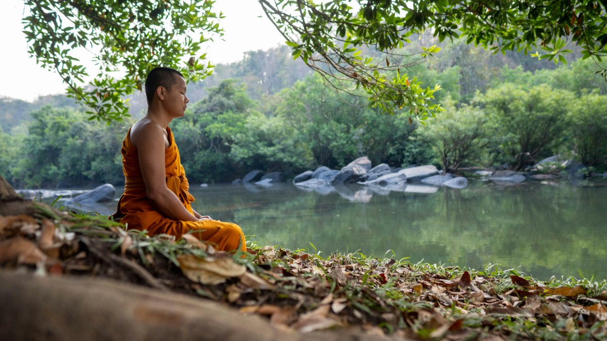 A person in orange robes sits meditating by a tranquil river, surrounded by lush greenery and rocks.