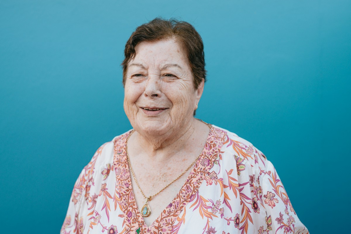 Elderly woman wearing a floral blouse stands against a blue background, looking ahead and smiling slightly.