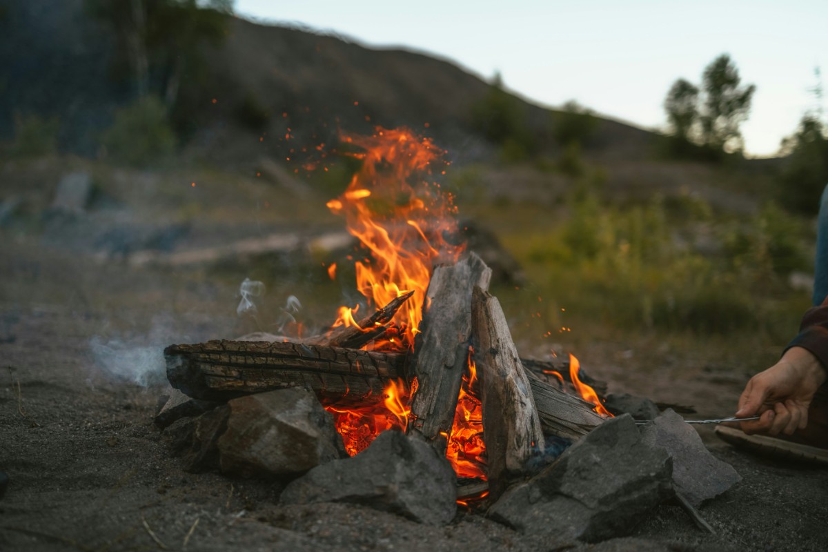 A campfire burning with logs and surrounded by rocks in an outdoor setting, with trees and a hill in the background.
