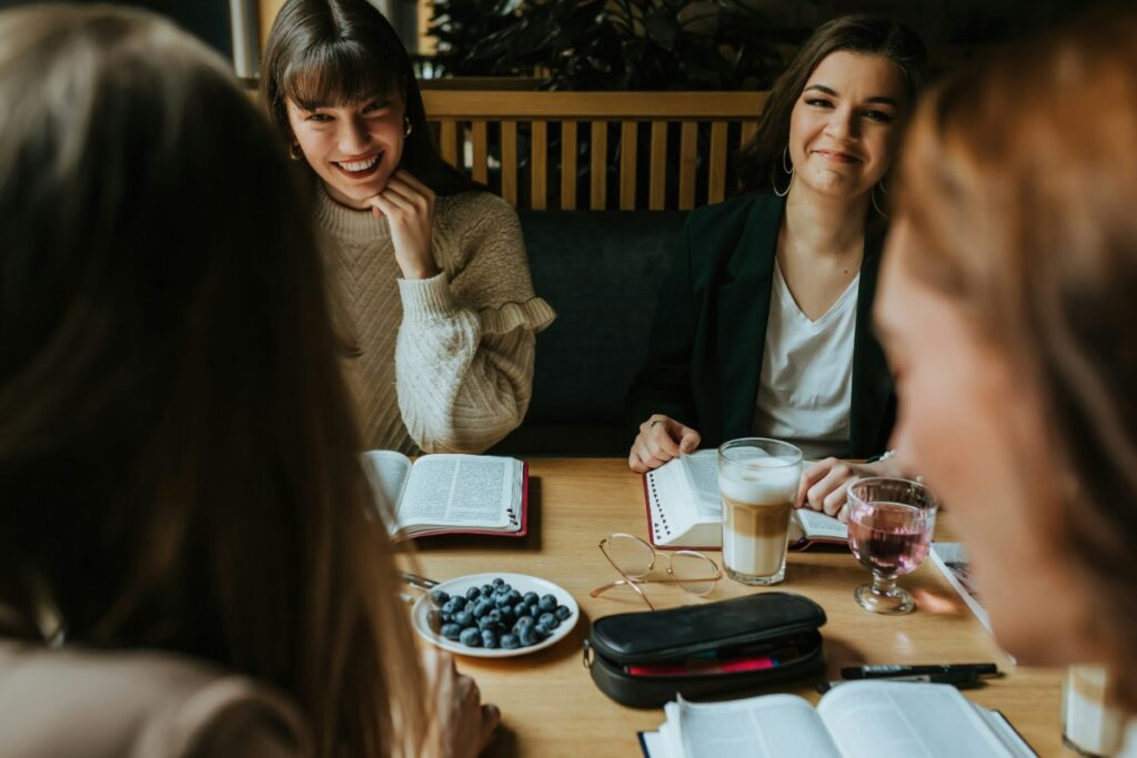 Four people sitting at a table with open books, a glass of wine, and a bowl of blueberries.