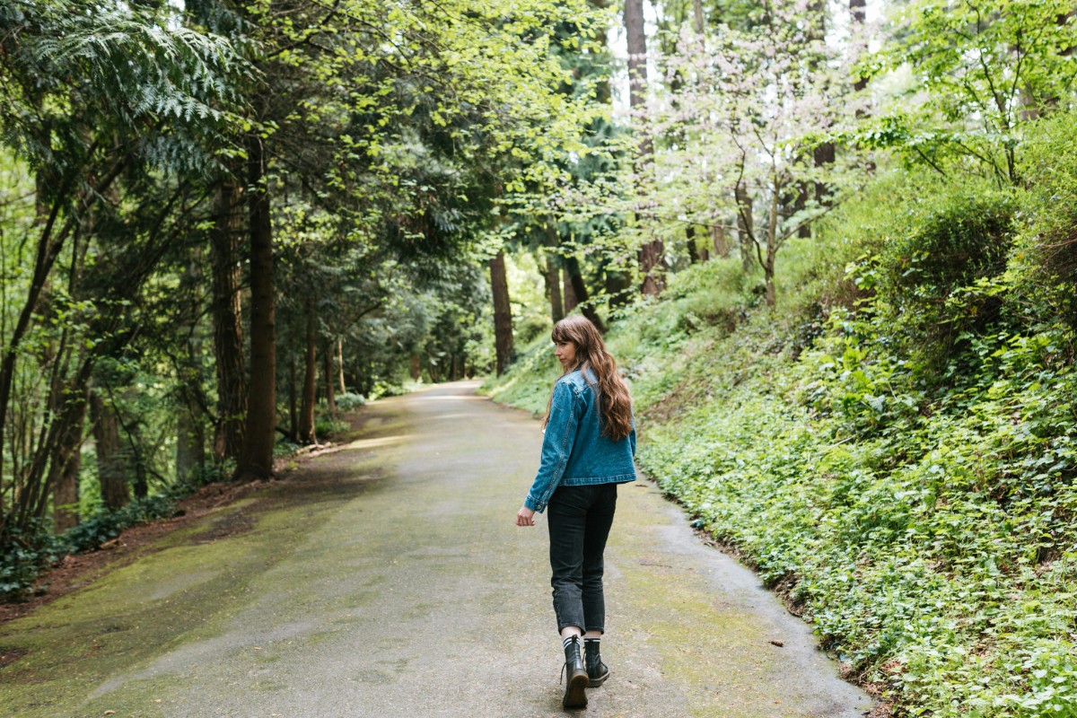 A person with long hair walks on a forest path, surrounded by green trees and foliage.