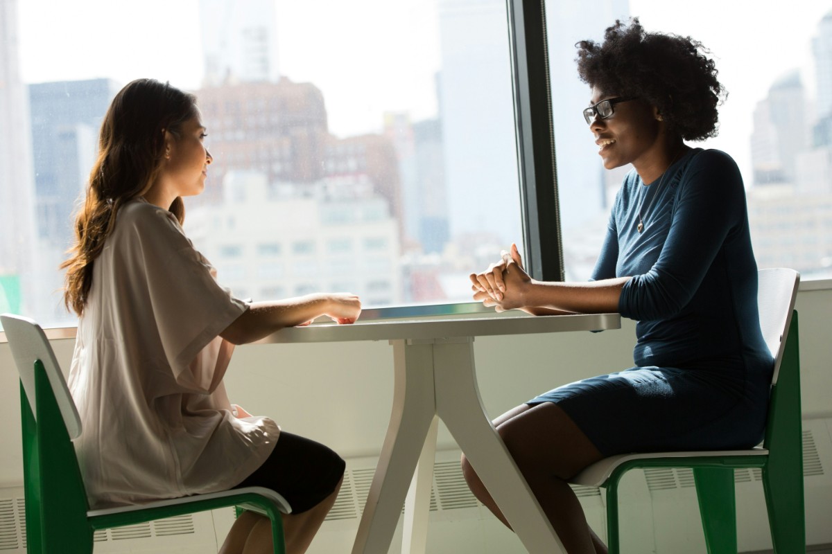 Two people sitting at a table by a window, engaged in conversation with a cityscape in the background.