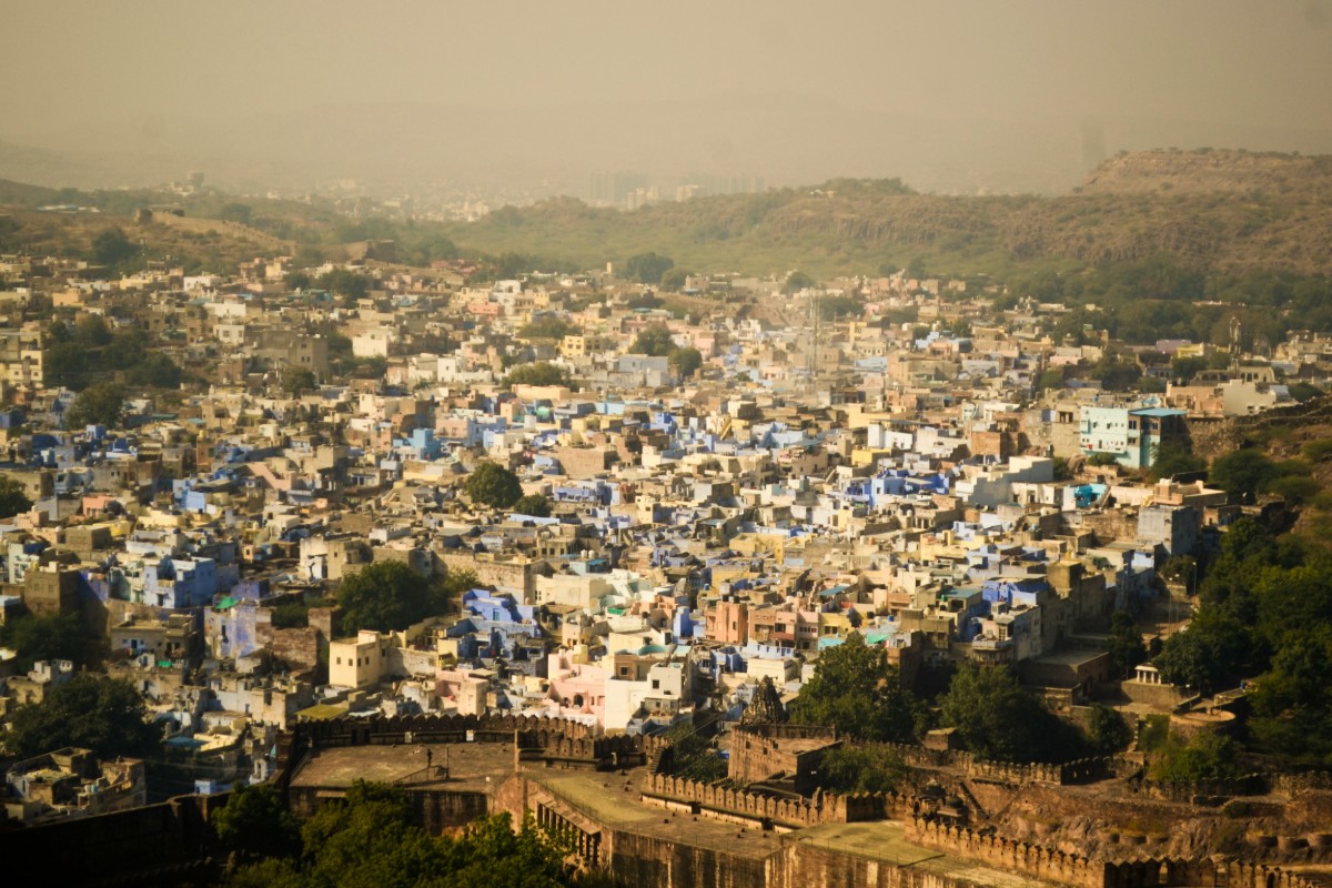 Aerial view of a densely packed cityscape with numerous blue-painted buildings, surrounded by trees and hills under a hazy sky.