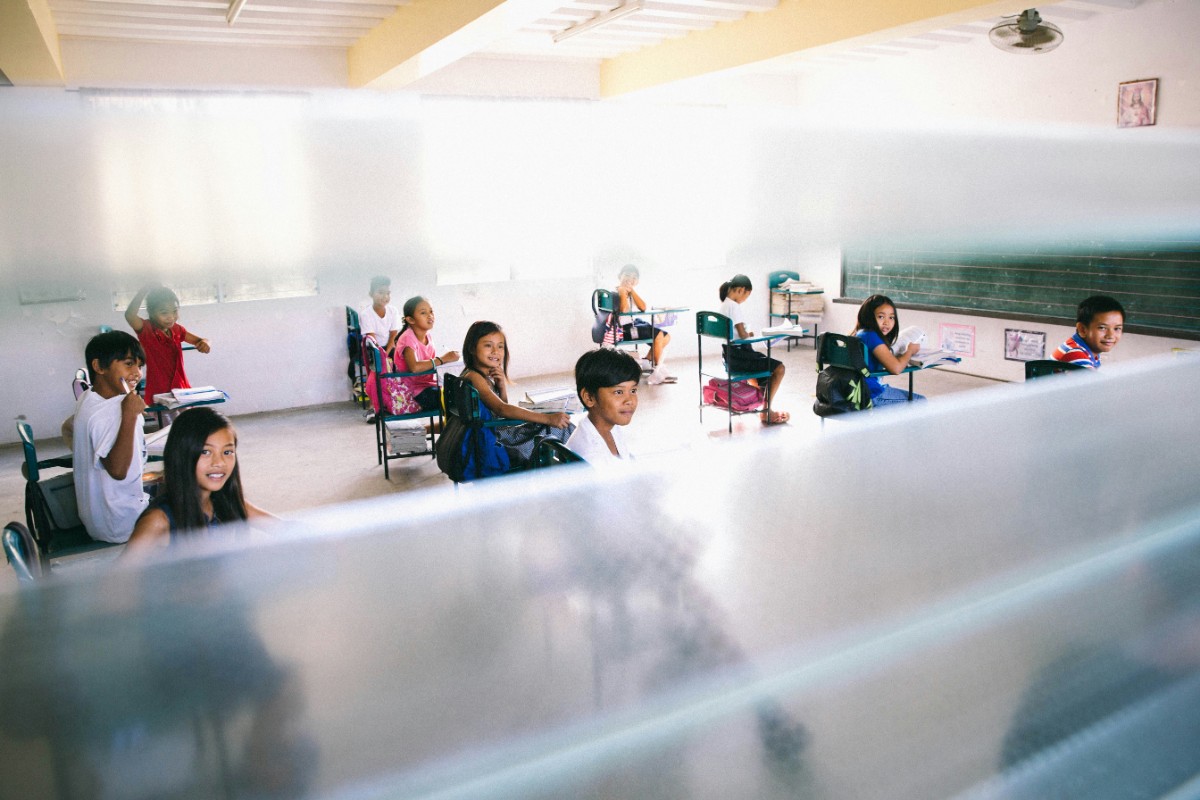 Students sitting at desks in a classroom, separated by transparent partitions, attentive to a lesson.