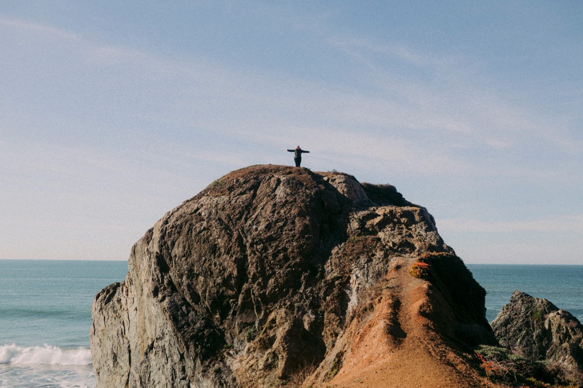 Person standing on a large rocky cliff overlooking the ocean under a clear blue sky.