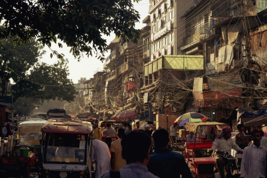 Busy street scene with people, auto rickshaws, and tangled overhead wires. Buildings line the street under a partly cloudy sky.