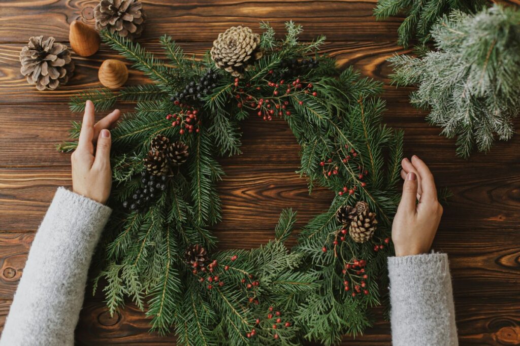 Hands arranging a festive wreath with pine cones and berries on a wooden surface, surrounded by additional pine cones and greenery.