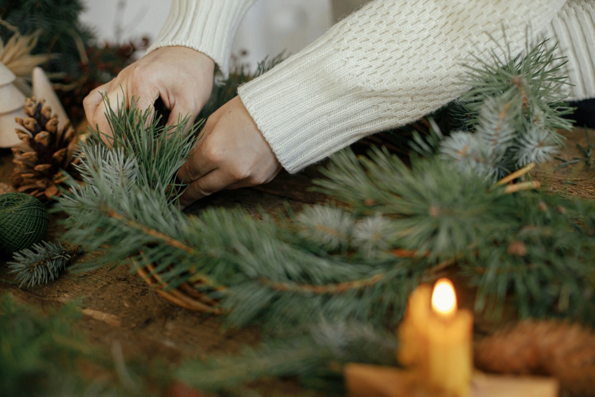 Hands arranging a pine garland on a table, with a lit candle nearby.