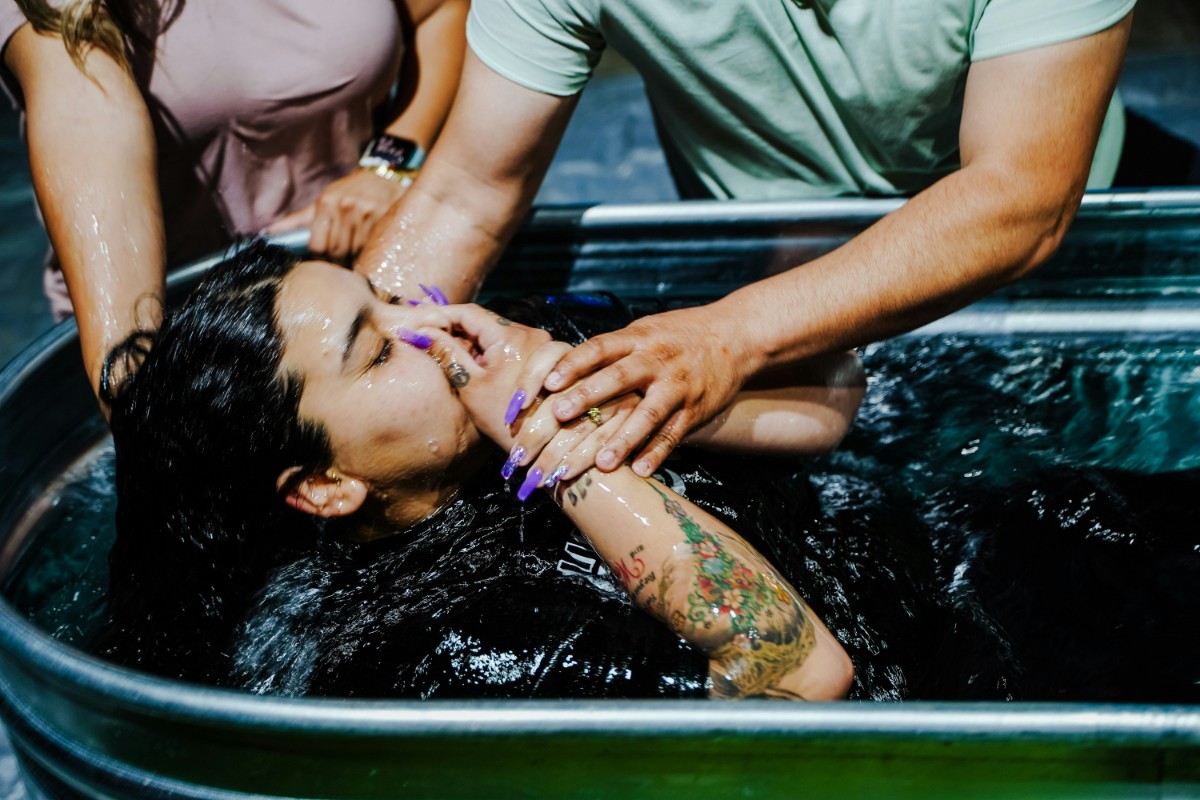 Person being baptized by immersion in a metal tub, assisted by two others.