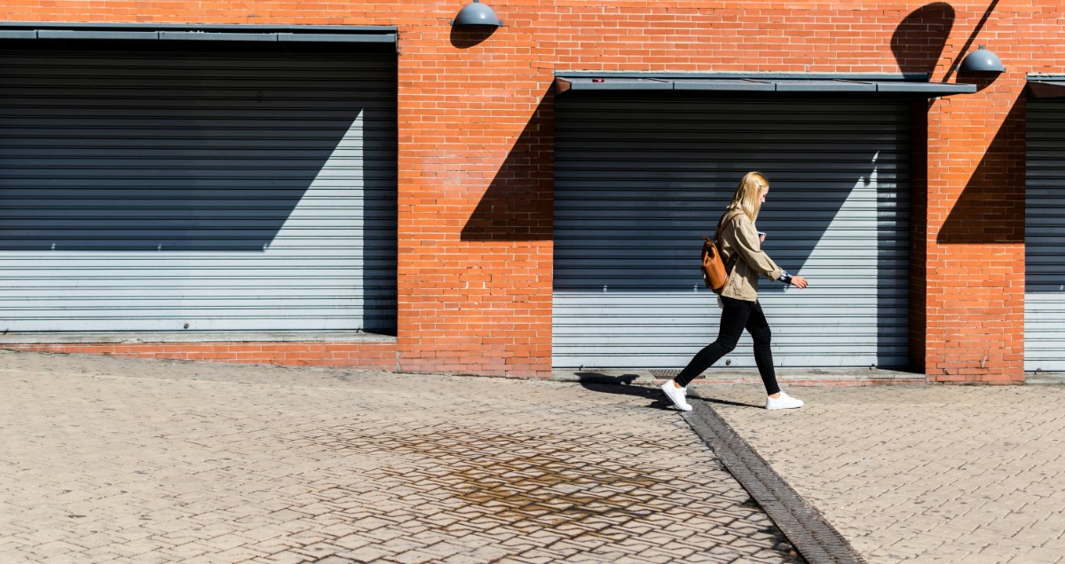 A person wearing casual clothes walks along a paved area in front of a brick building with three closed garage doors.