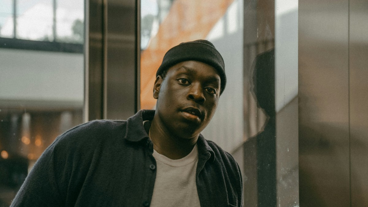 A person wearing a beanie and a dark jacket stands indoors, looking toward the camera. The background includes glass and abstract reflections.