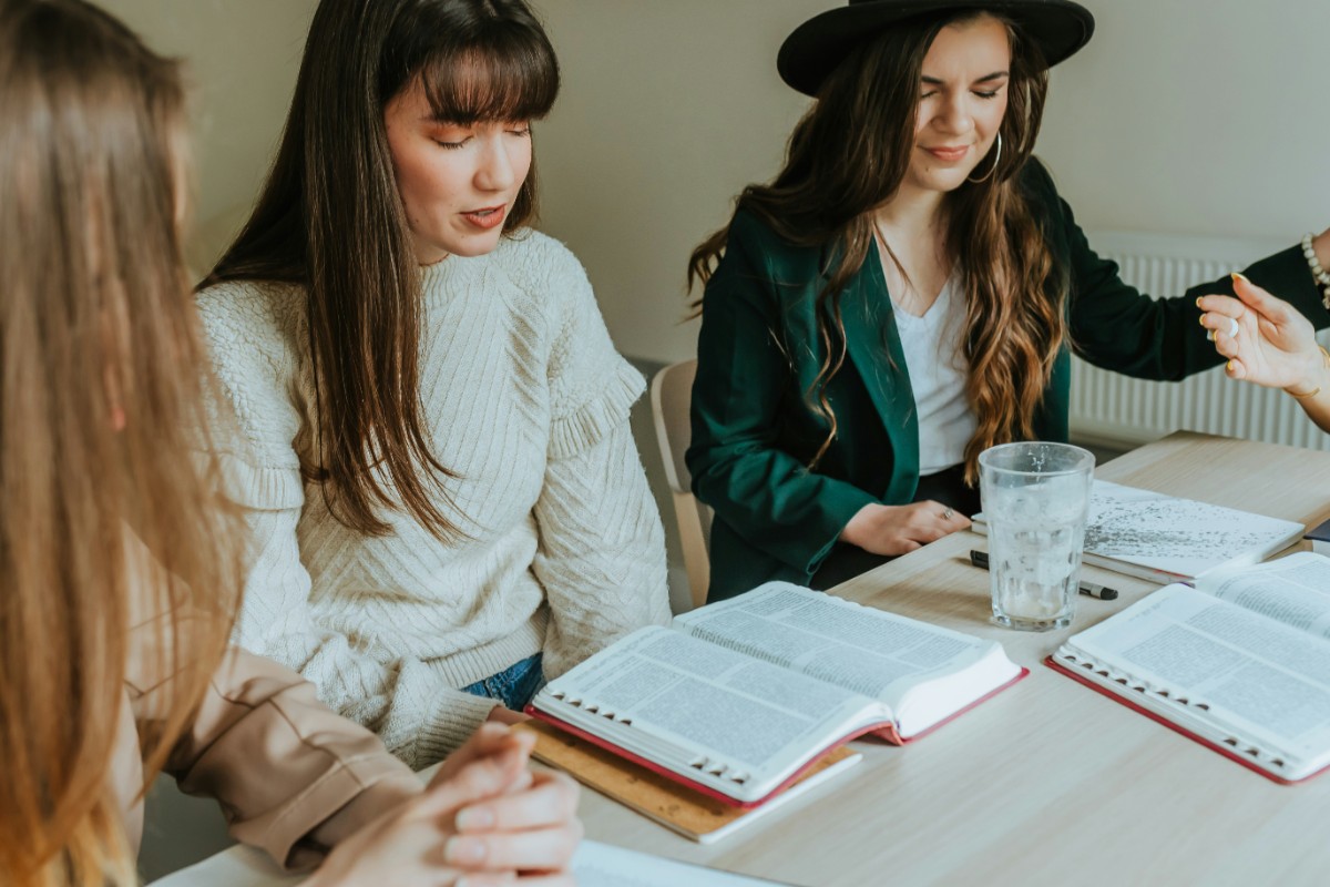 Three women are sitting at a table with open books, engaged in discussion. One woman is wearing a hat. A glass of water is on the table.
