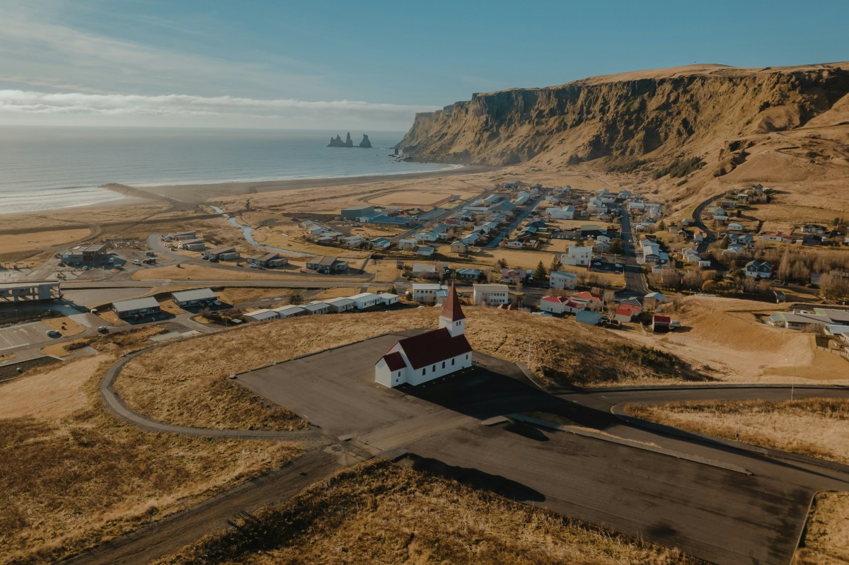 Aerial view of a coastal village with a red-roofed church in the foreground, cliffs in the background, and the ocean beyond. Houses are spread out across the landscape.
