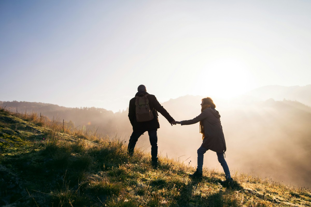 Two people hiking on a grassy hillside, with one reaching back to hold the others hand. The sun shines brightly in the background.