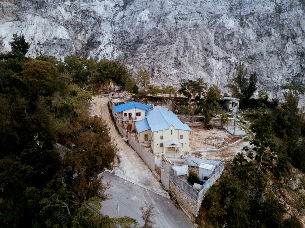 Small complex with buildings having blue roofs, surrounded by a wall and trees, situated near a rocky mountain slope.