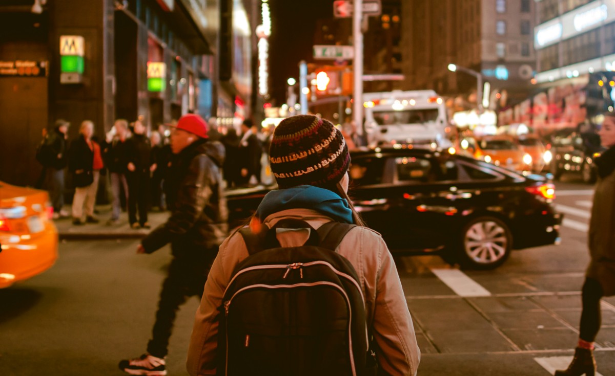 Person wearing a backpack stands at a busy city street corner at night, surrounded by cars and pedestrians.
