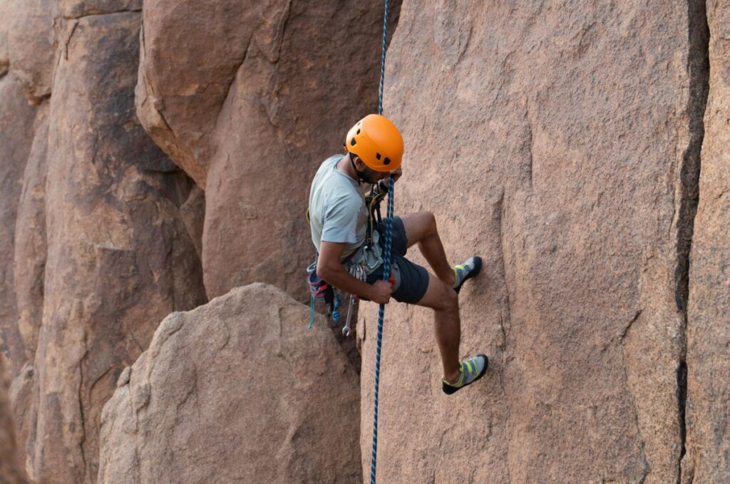 A climber wearing a helmet ascends a steep rock face using ropes and climbing gear.