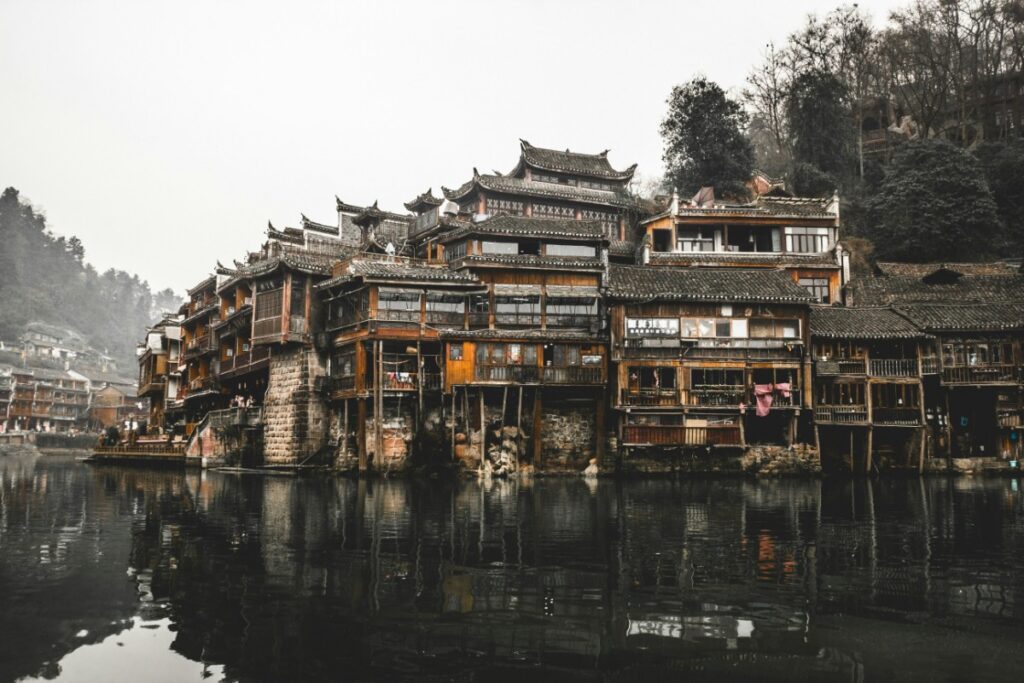 Traditional wooden buildings line a riverbank, reflected in the calm water, with trees and a foggy sky in the background.