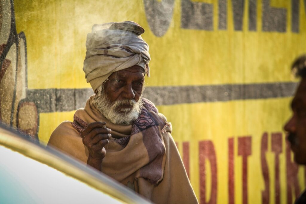 A man with a turban and beard stands against a yellow wall in sunlight, holding an object.