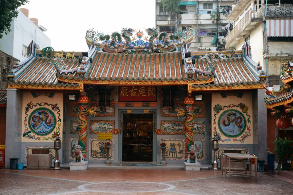Ornate traditional temple with colorful decorations, intricate carvings, and tiled roof. Surrounded by urban buildings and a brick courtyard in the foreground.