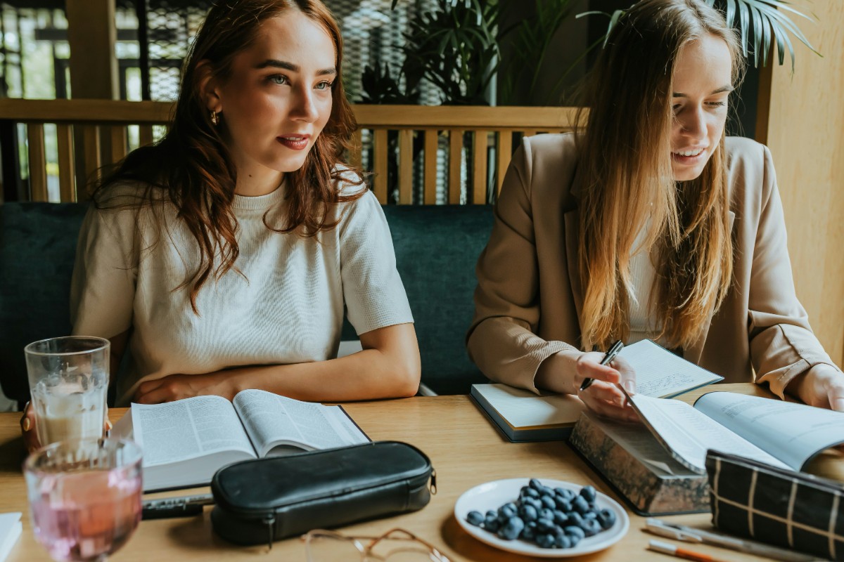 Two women sitting at a table, reading books. A pencil case and a plate of blueberries are on the table.