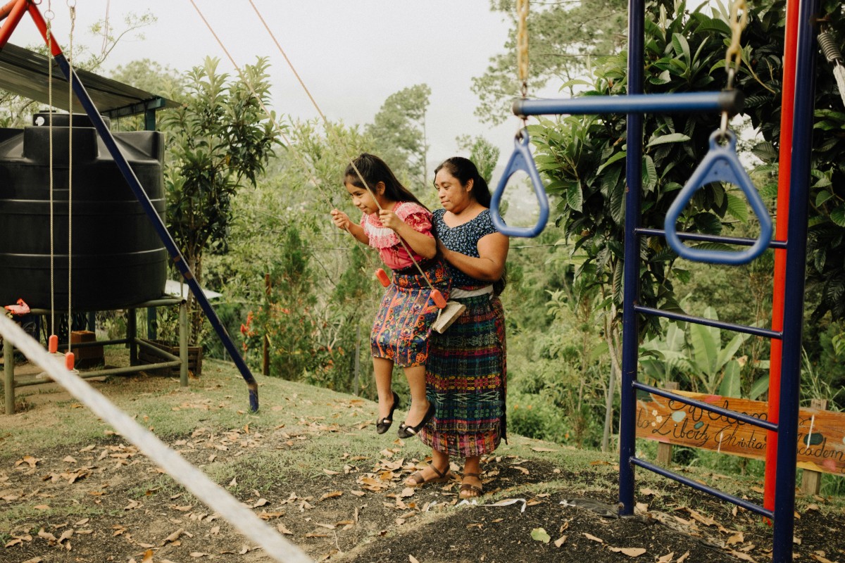 Two people on a playground, one adult helping a child in a swing set. Trees and greenery are in the background.