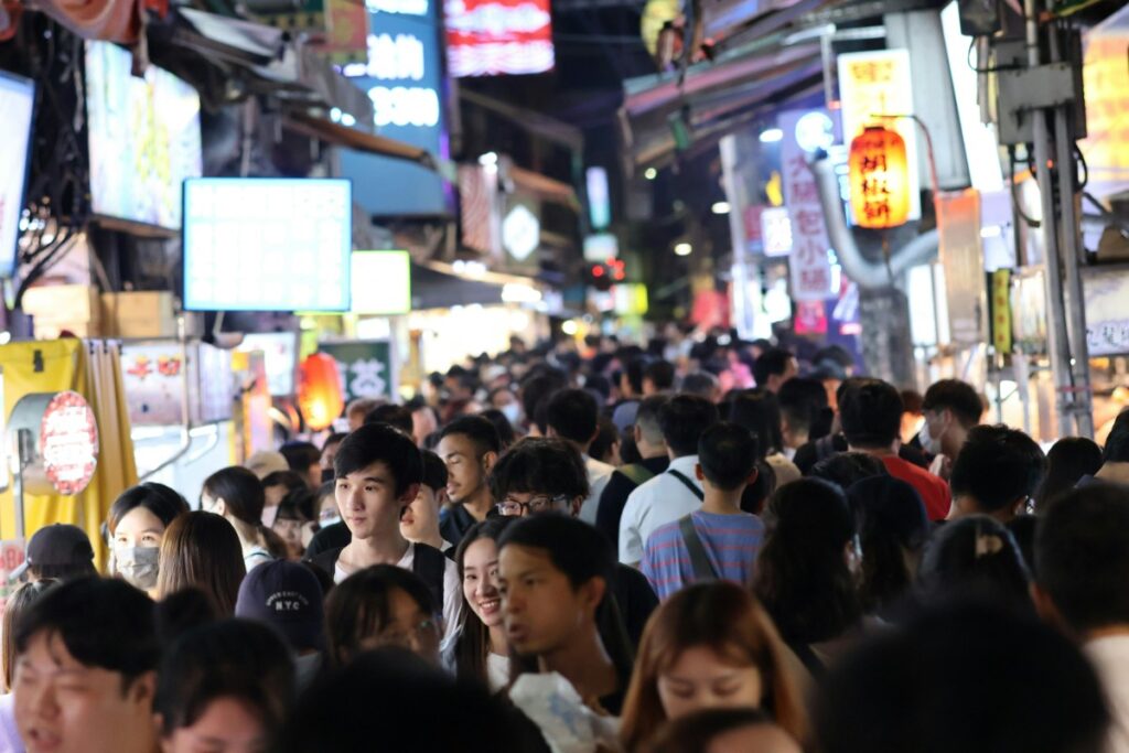 A large crowd of people walks through a busy night market, with bright signs and food stalls lining the narrow pathway.