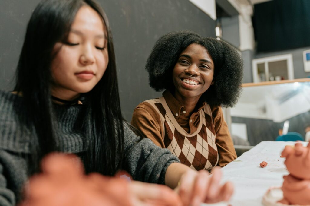Two people sitting at a table, engaged in a creative activity with clay. One person is smiling while looking at the other.
